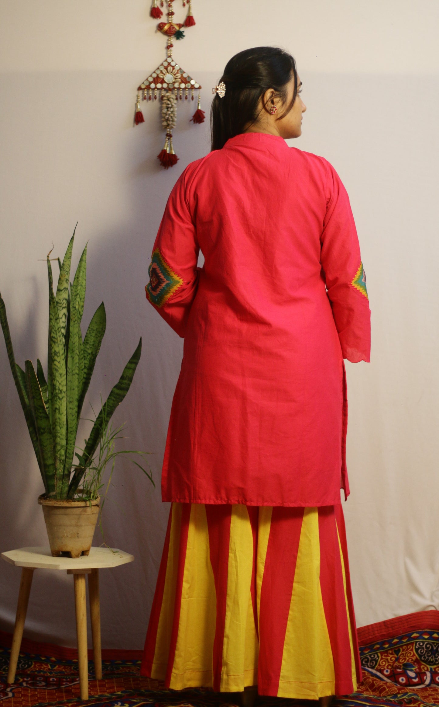 Person wearing a red kurta with yellow pants standing against a white wall.