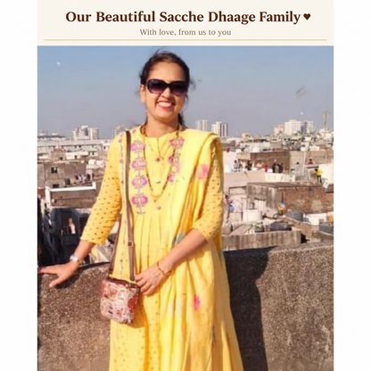 A woman wearing a bright yellow embroidered kurta with pink floral details, sunglasses, and a patterned crossbody bag, standing on a rooftop with a city skyline in the background.
