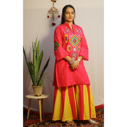 Woman wearing a bright pink embroidered top and yellow pants standing indoors with a plant and decorative items in the background.