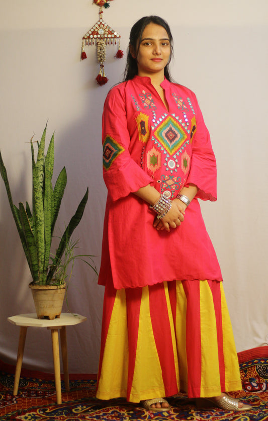 Woman wearing a bright pink embroidered top and yellow pants standing indoors with a plant and decorative items in the background.